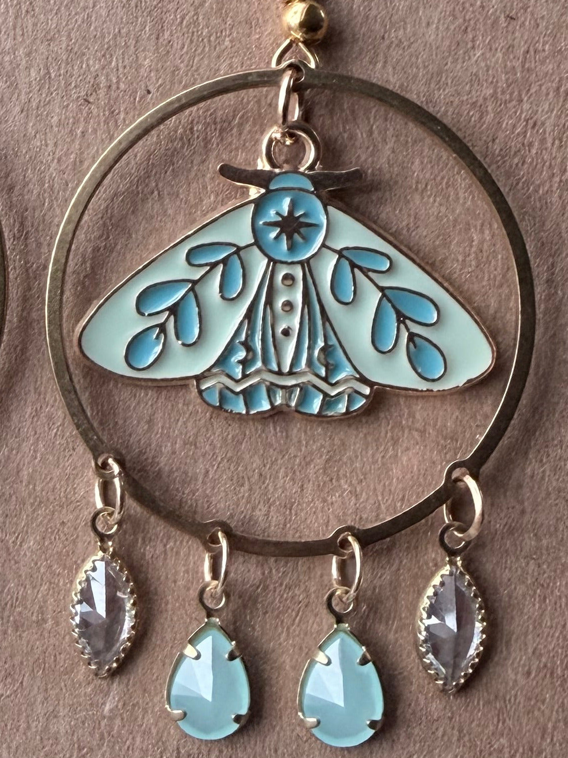 old-tone dangle earring displayed on a brown card. Each earring has a circular hoop with a butterfly motif in light blue and turquoise enamel, a central blue stone, and three dangling teardrop charms including one light blue stone and two silver-toned stones.