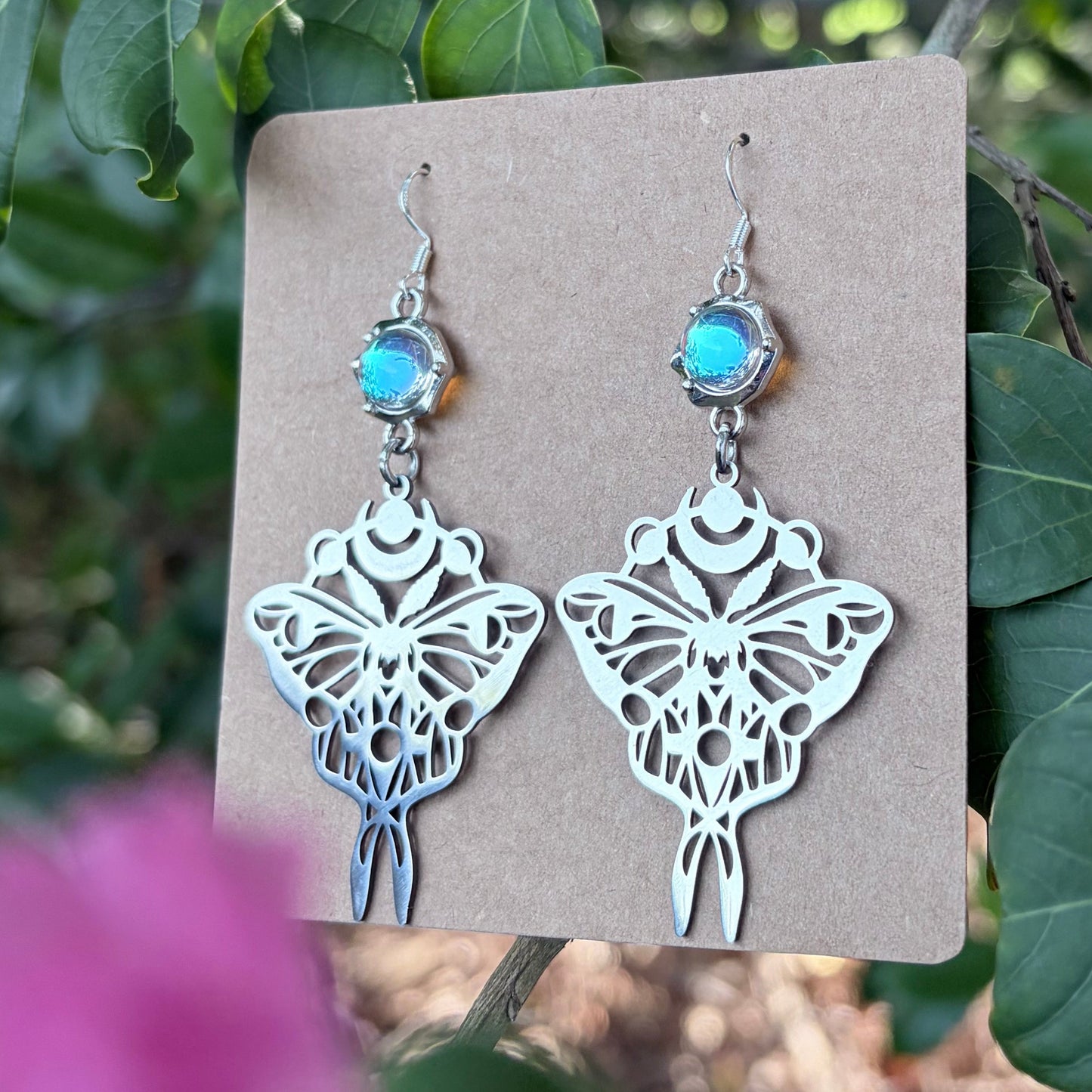 Silver-toned moth dangle earrings with small turquoise-blue stones, displayed on a kraft card against a background of green leaves and blurred pink flowers.