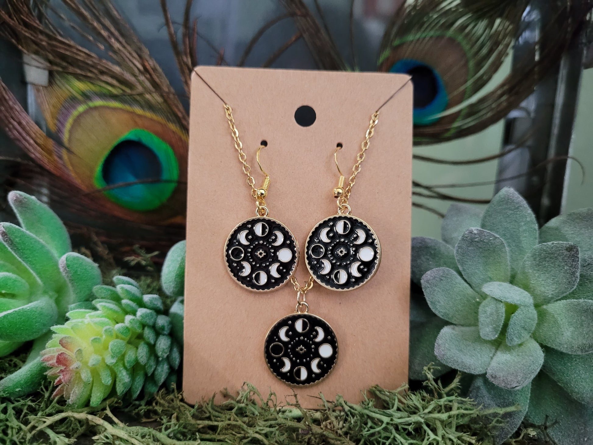 Pair of black and gold earrings on a beige card with peacock feathers and succulents in the background