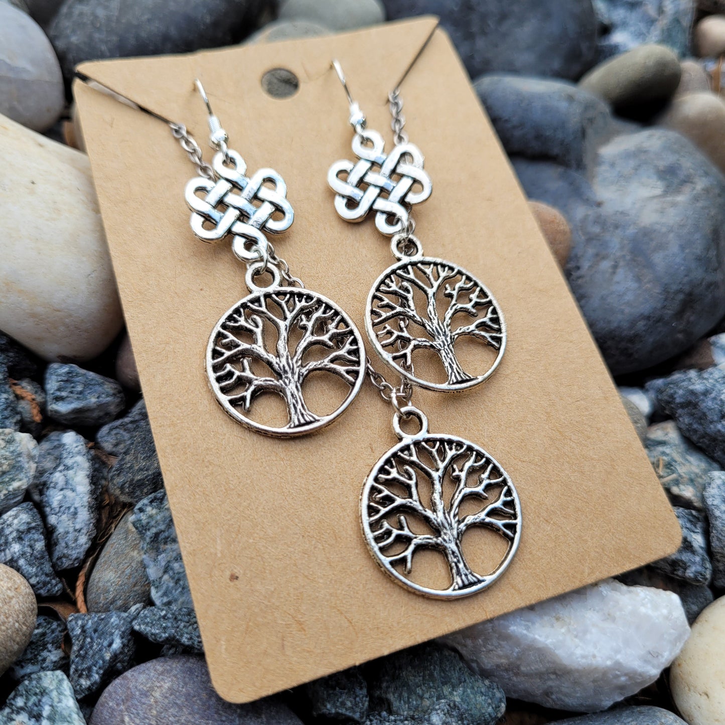 Silver tree-shaped earrings on a cardboard backing with pebbles in the background