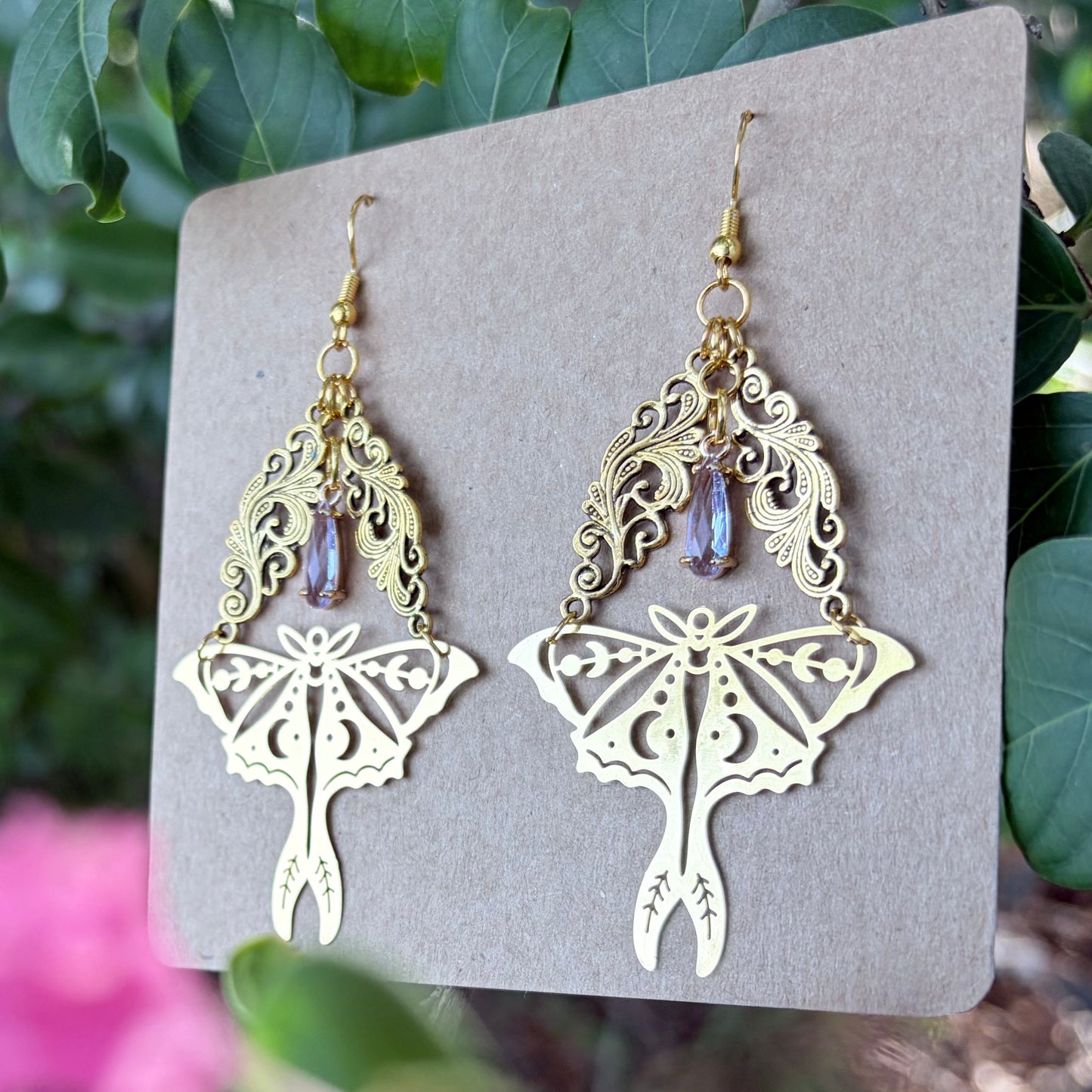 A pair of gold-toned dangling earrings displayed on a square kraft card, photographed outdoors against green leaves and blurred pink flowers.