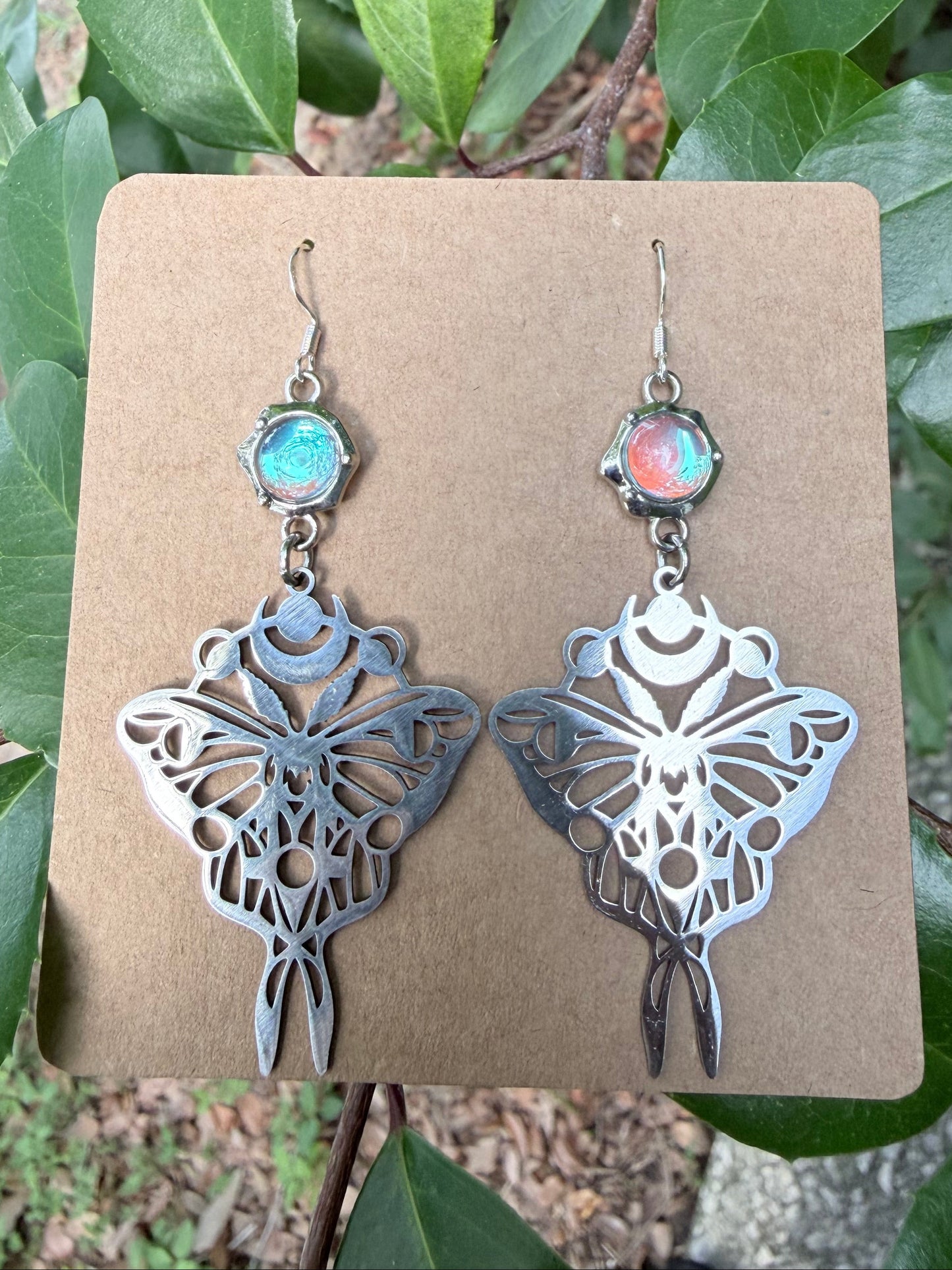 A pair of dangling luna moth-shaped earrings displayed on a brown card, photographed outdoors against green leaves. Each earring has a silver-toned moth cutout with an iridescent oval stone above it, one showing a blue-green sheen and the other a pink-orange sheen.
