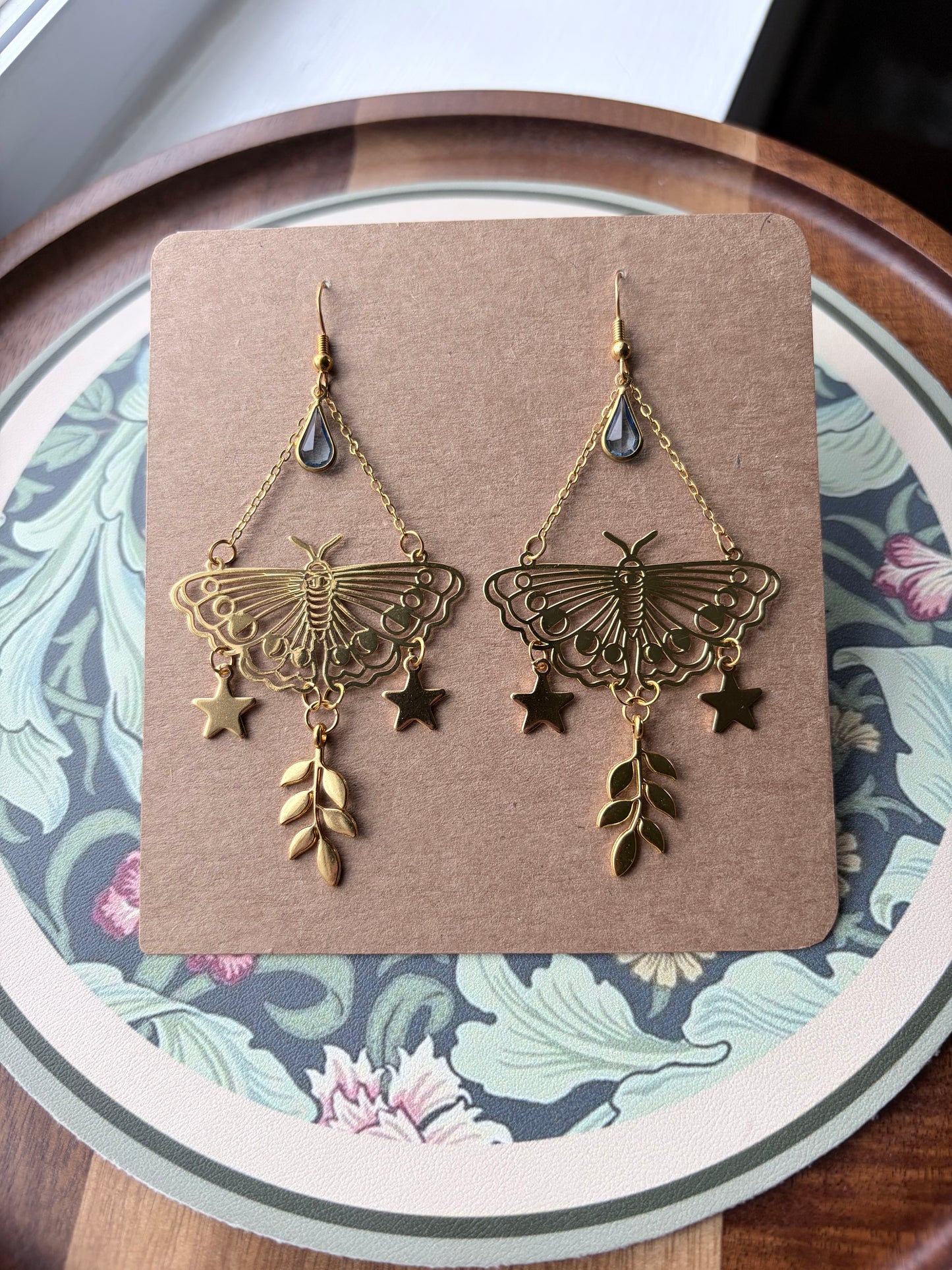 A pair of dangling earrings displayed on a brown card, resting on a decorative floral plate. The earrings are made of gold-toned metal with a butterfly motif, small star charms, and leaf charms, with a small dark teardrop stone at the top.