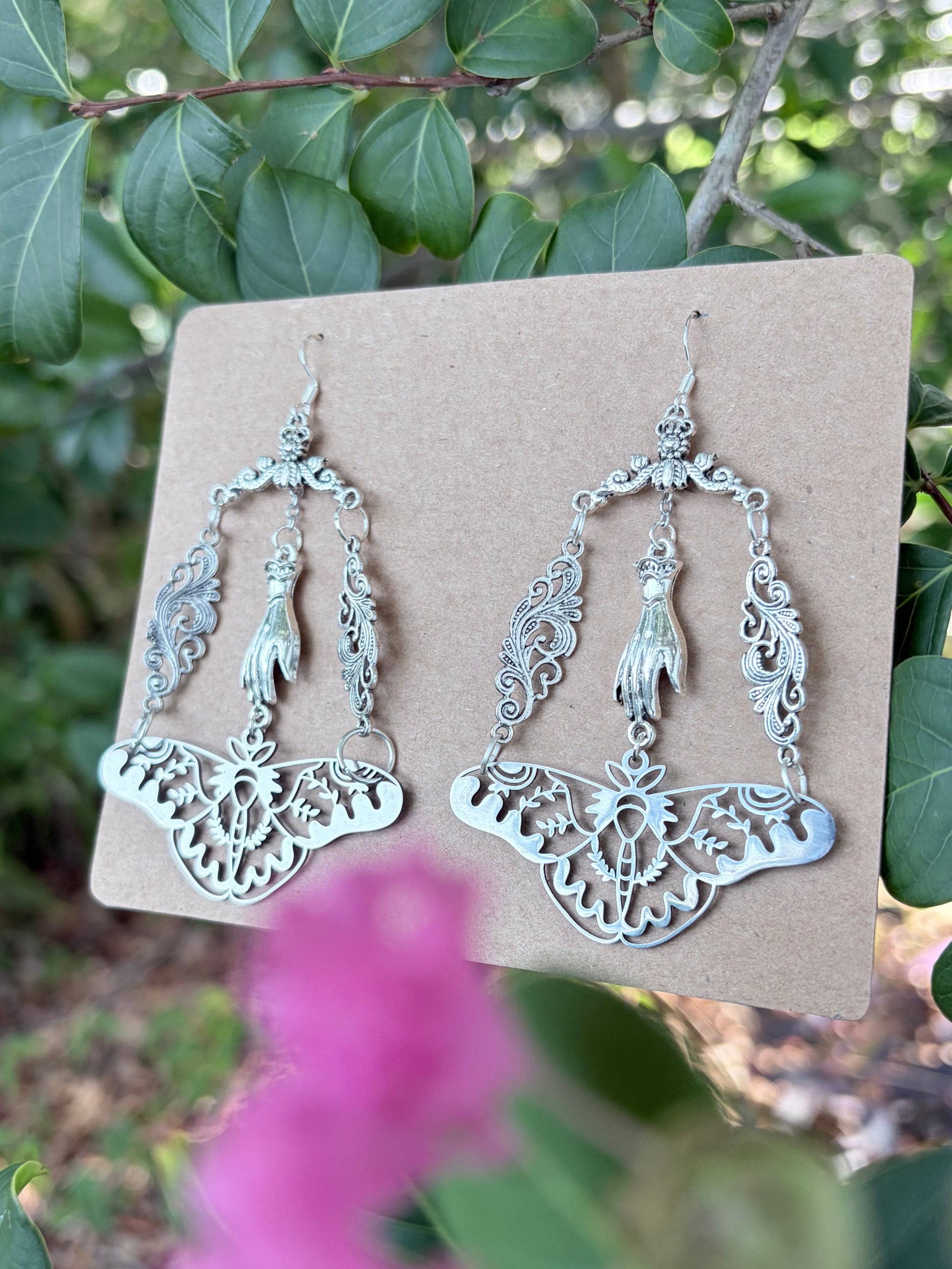 Silver-toned filigree dangle earrings displayed on a tan card, photographed outdoors against green leaves with a blurred pink flower in the foreground.