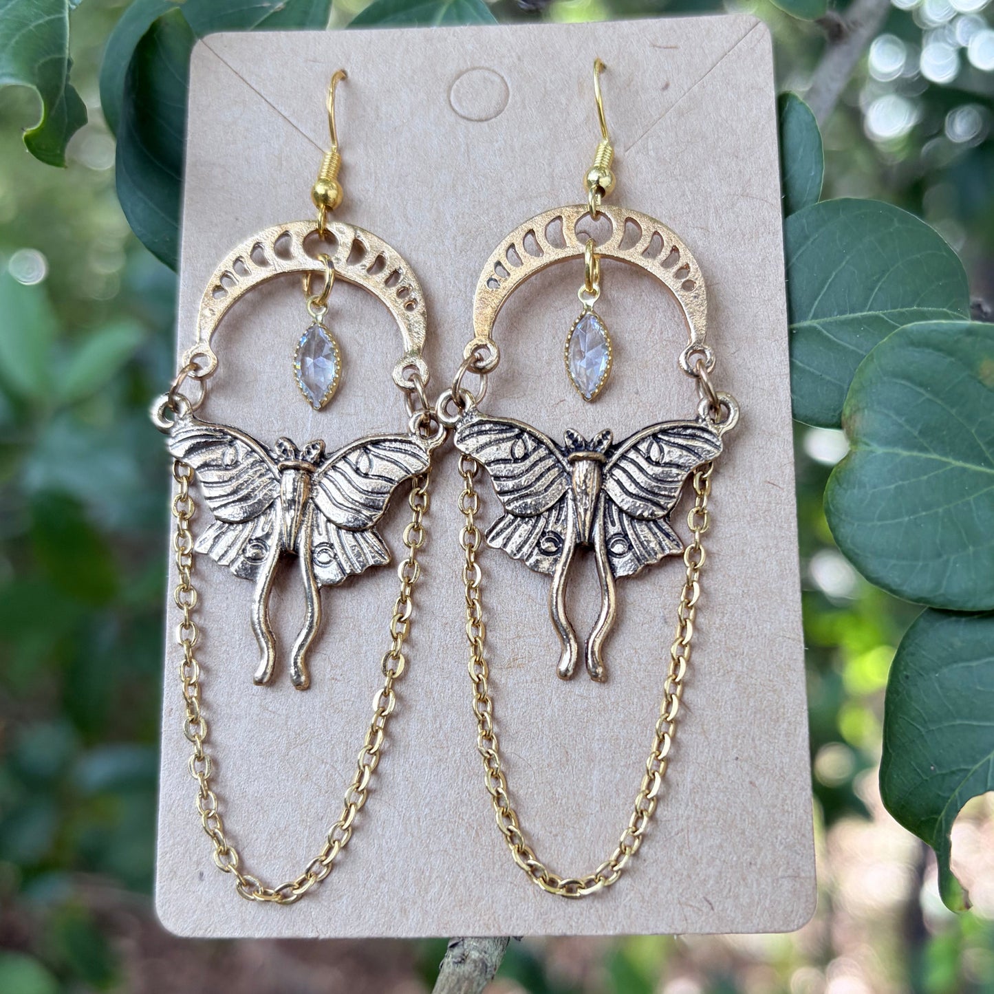 A pair of dangling earrings displayed on a small beige card, photographed outdoors against green leaves and branches. Each earring has a gold-tone crescent moon and butterfly motif with a small clear teardrop stone and a longer chain hanging below.