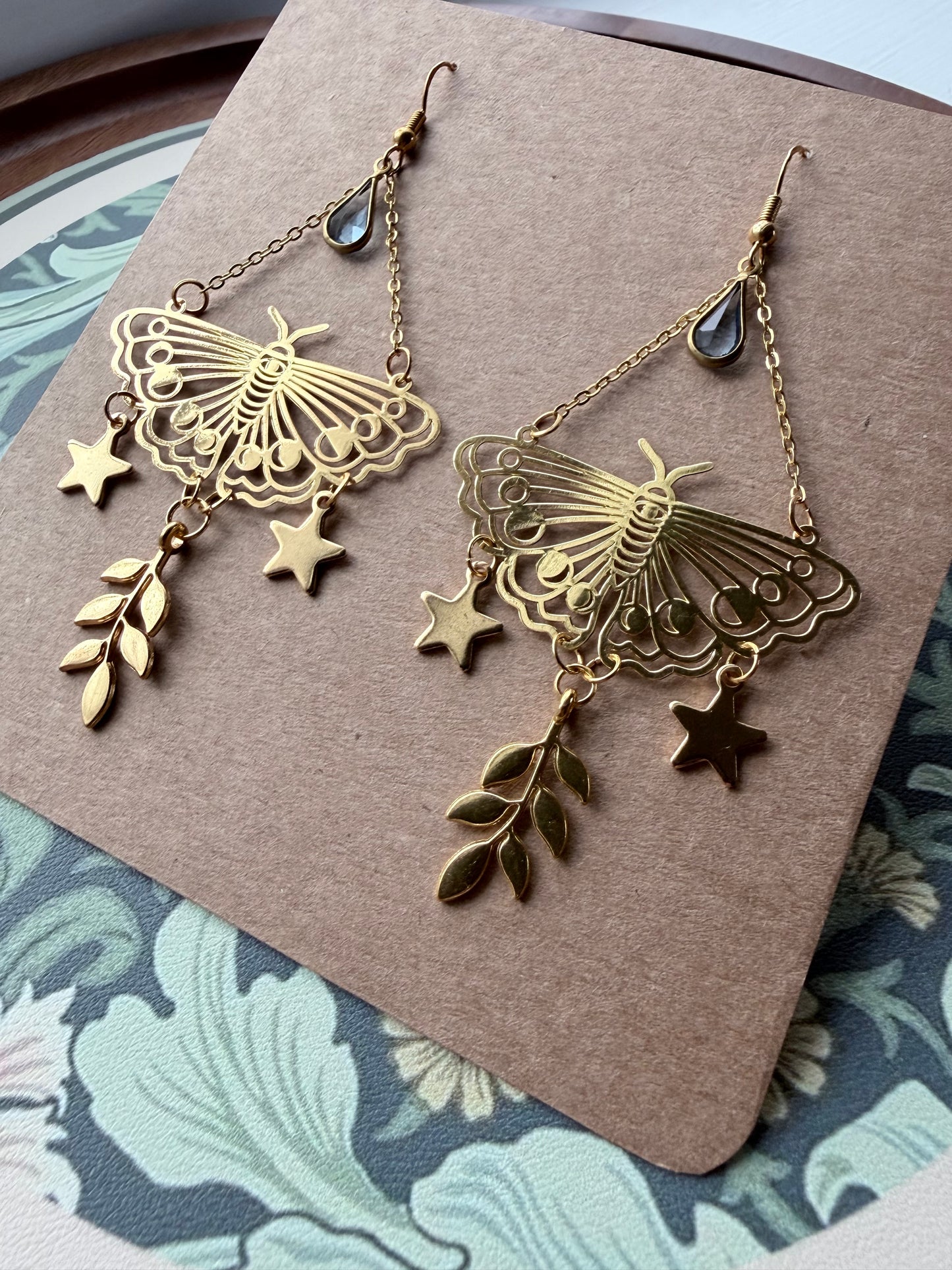 A pair of gold-toned dangling earrings displayed on a brown kraft card. Each earring features a butterfly-shaped filigree charm, small star charms, and leaf charms, with a dark teardrop bead near the top.