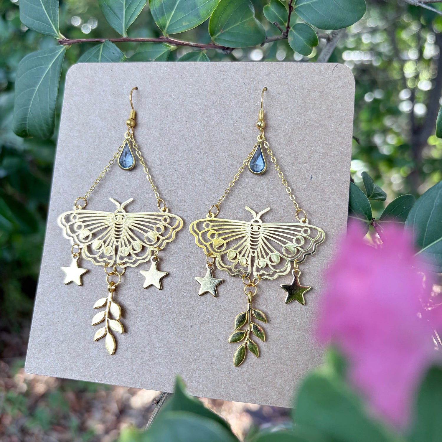 A pair of gold-toned dangle earrings displayed on a flat cardboard card, photographed outdoors with green leaves and a pink flower in the blurred background. Each earring has a butterfly-shaped charm, small star charms, a leaf charm, and a small blue teardrop bead near the top.