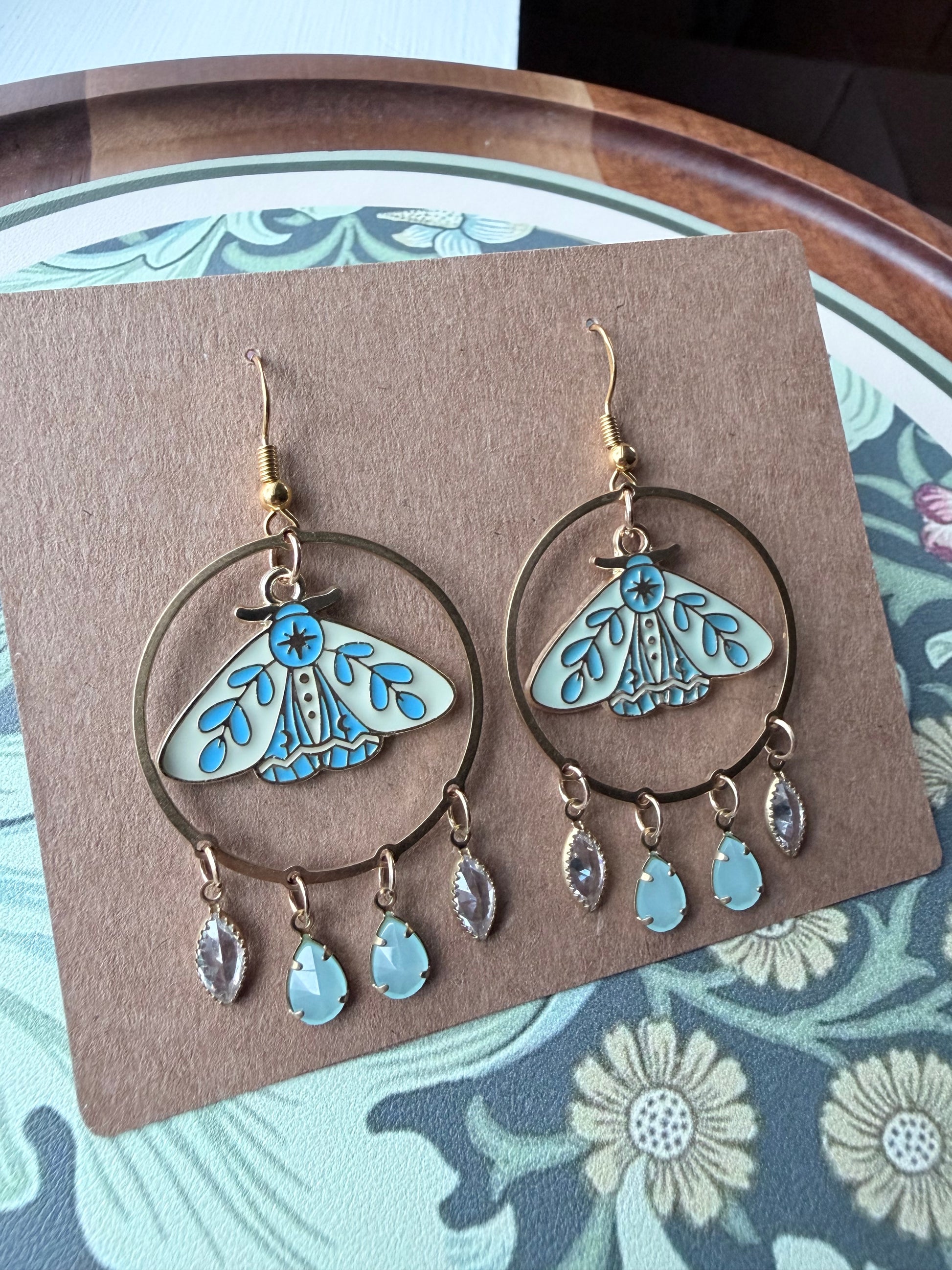 A pair of dangling hoop earrings displayed on a brown card. Each earring has a large gold-tone hoop with a light blue and turquoise moth design in the center and three small dangling teardrop charms below.