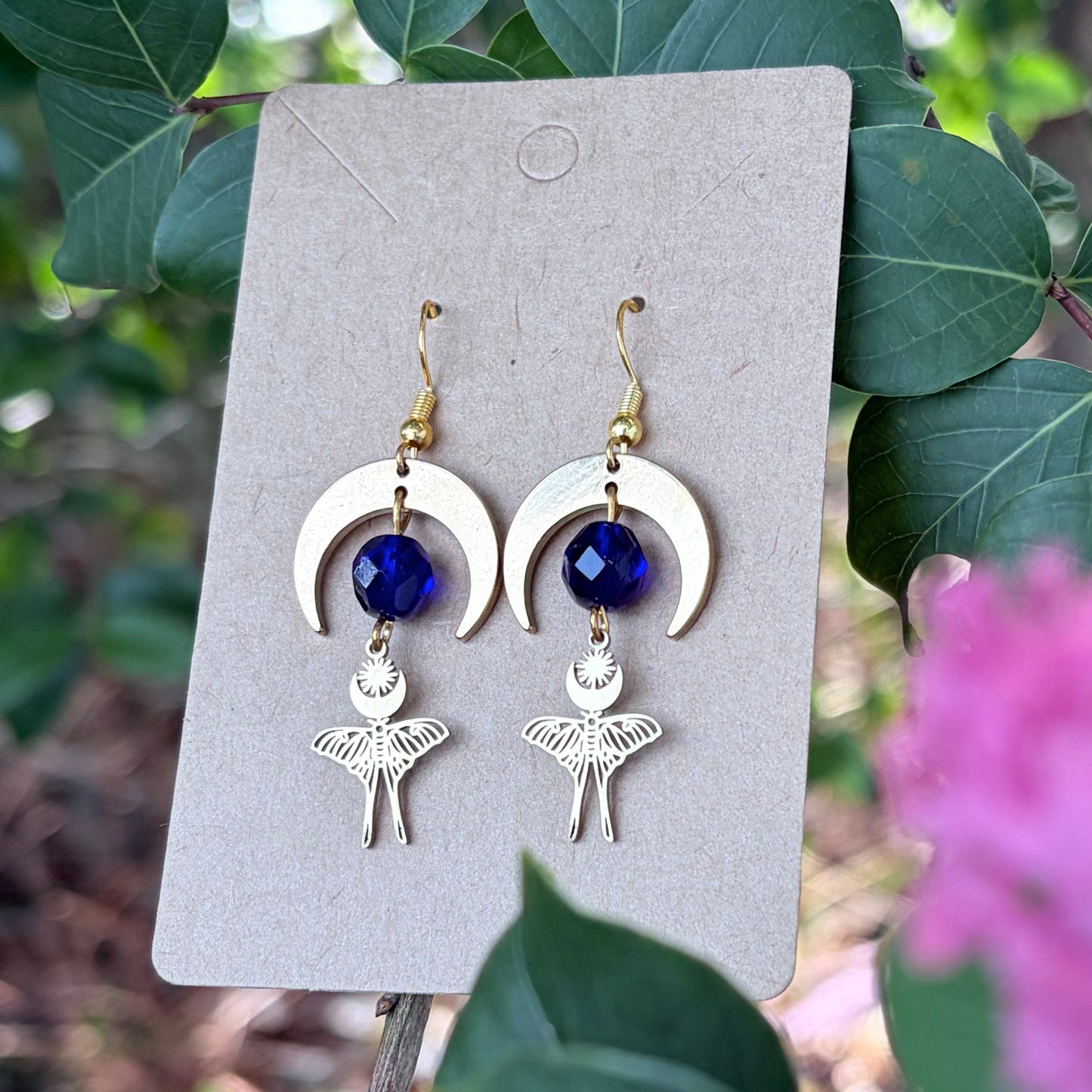 A pair of handmade dangle earrings displayed on a small beige card, set against a natural background of green leaves and pink flowers. The earrings feature gold-toned crescent moon and luna moth charms with deep blue faceted beads.