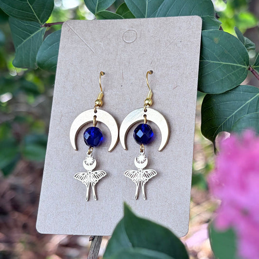 A pair of handmade dangle earrings displayed on a small beige card, set against a natural background of green leaves and pink flowers. The earrings feature gold-toned crescent moon and luna moth charms with deep blue faceted beads.