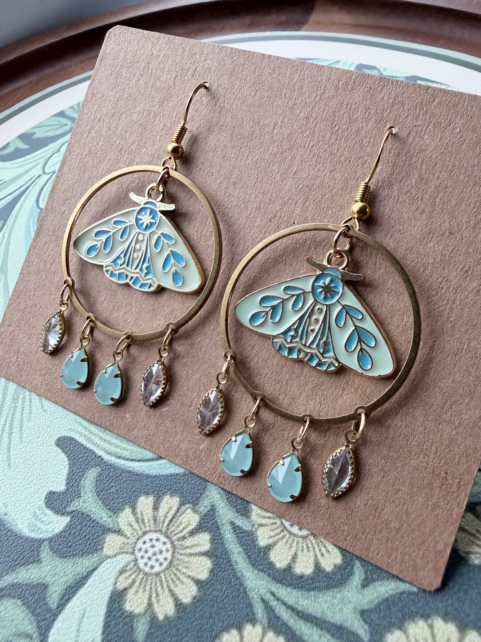 A pair of dangling hoop earrings displayed on a brown card. Each earring has a large gold-tone hoop with a light blue and white butterfly motif in the center and three dangling teardrop charms, including one light blue stone and two clear faceted stones.